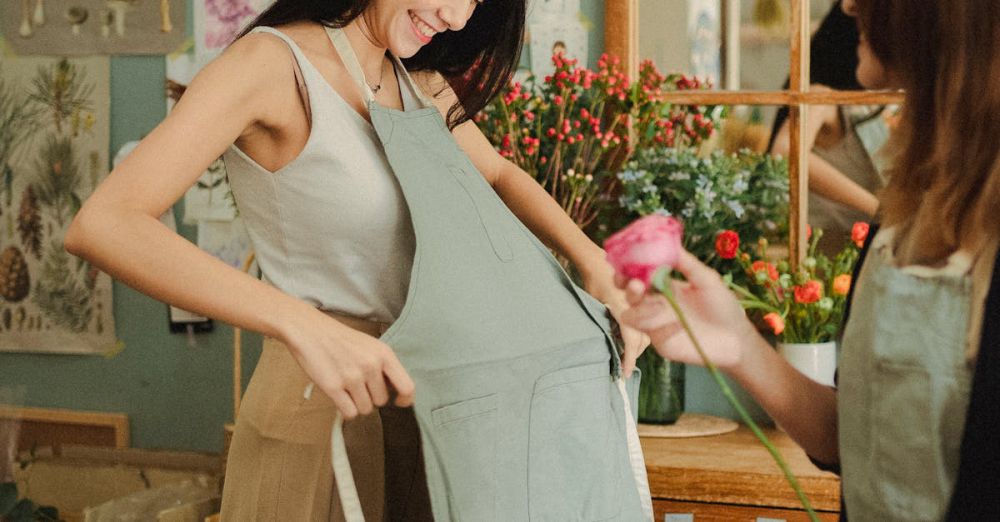 Craft Market Tour - Positive young woman putting on apron while preparing for work in floral shop with coworker